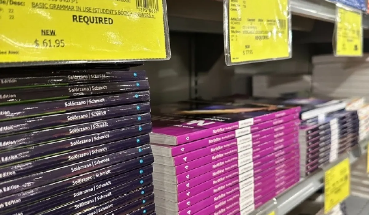 Shelves stacked with textbooks in various colors, including purple, red, and blue, with bright yellow price and course requirement tags displayed on the edge of the shelves.