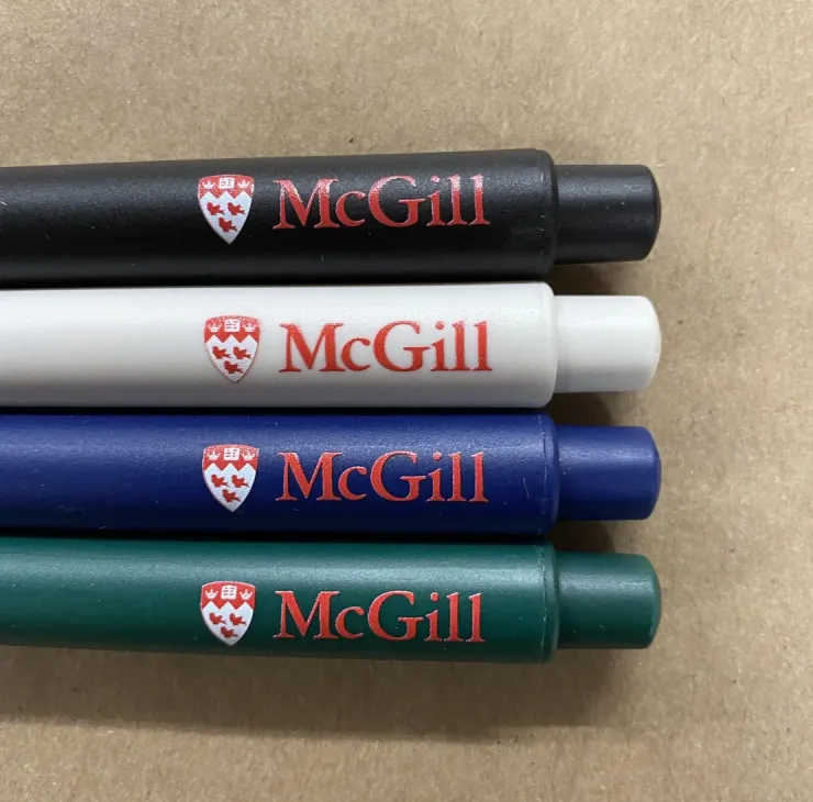 Set of four McGill-branded recycled pens in green, blue, black, and white, made in Canada, displayed on wooden surface.