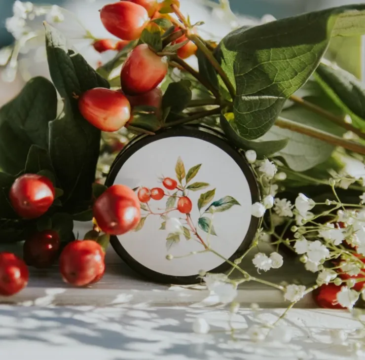 The Plains body balm in black tin with botanical rosehip illustration, displayed among red berries and greenery.