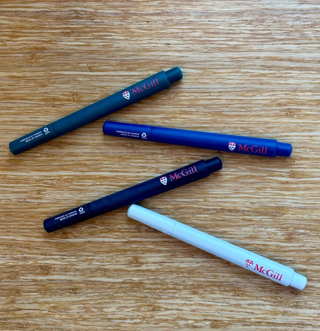 Set of four McGill-branded recycled pens in green, blue, black, and white, made in Canada, displayed on wooden surface.