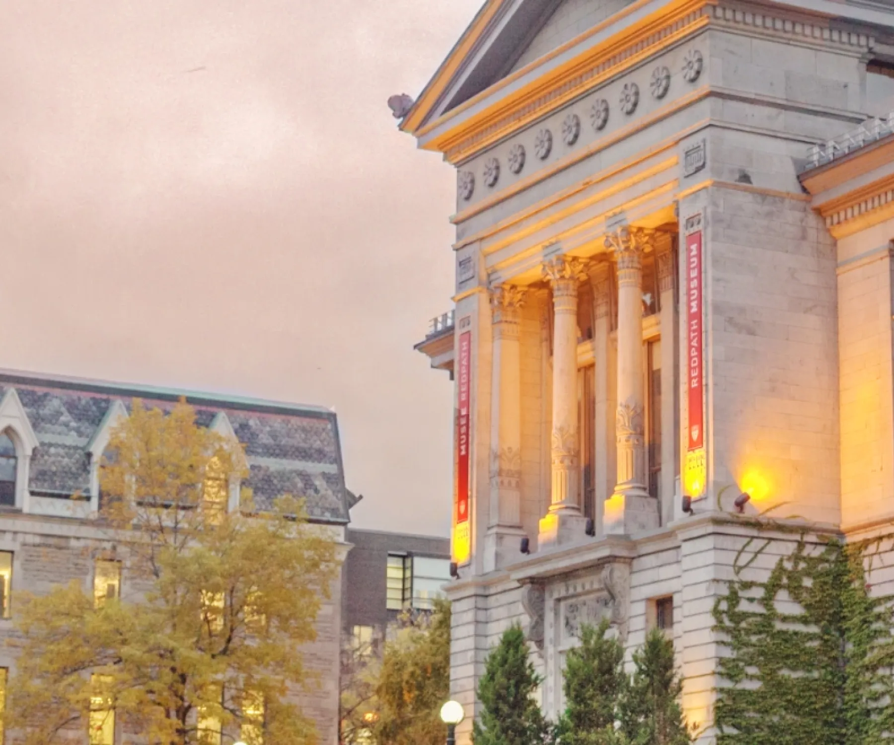 Exterior view of McGill University’s Redpath Museum and adjacent historic building during autumn. The scene shows warm evening lighting on the museum’s columns, colorful fall foliage, bicycles lined up along the sidewalk, and several people walking on the campus path