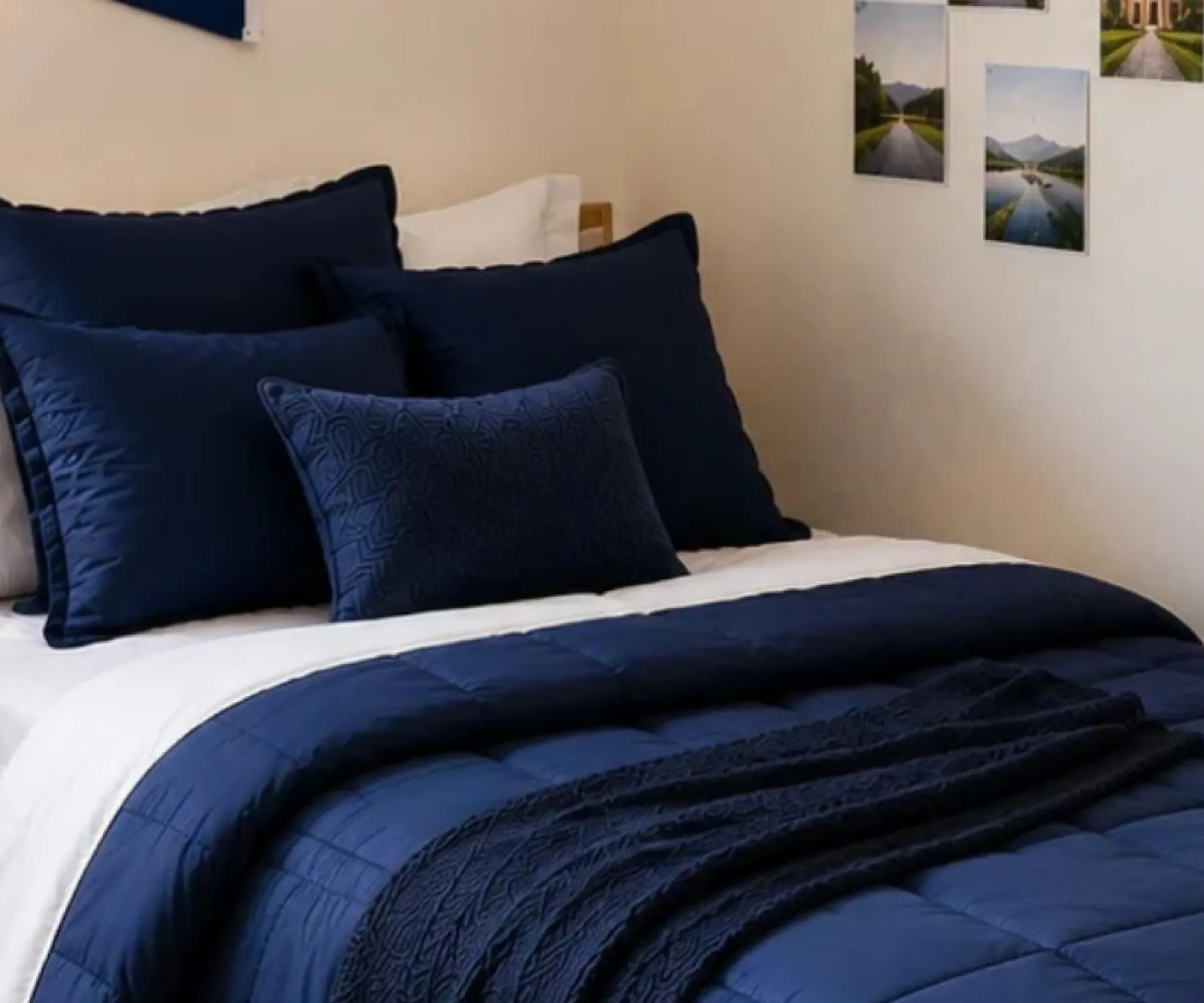 Neatly made bed with navy bedding and pillows in a bright dorm room, with plants, lamp, desk, and window.