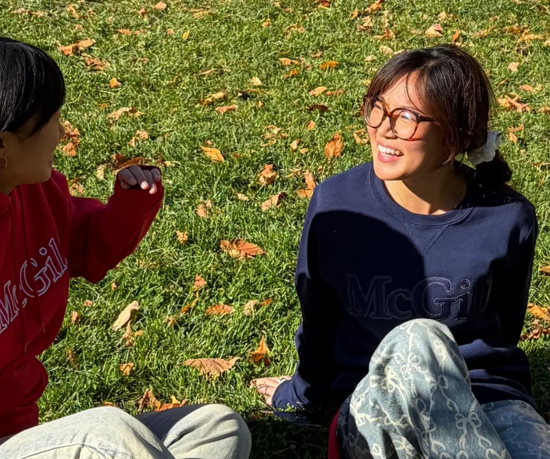 Two students wearing McGill sweaters sit on a sunny campus lawn scattered with colourful fall leaves.