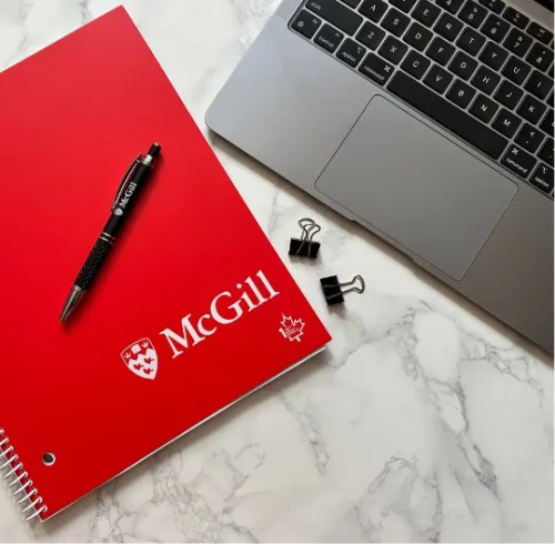 Flat lay of a workspace with a red McGill spiral notebook and black McGill pen on a white marble surface, next to a silver laptop keyboard and three black binder clips.